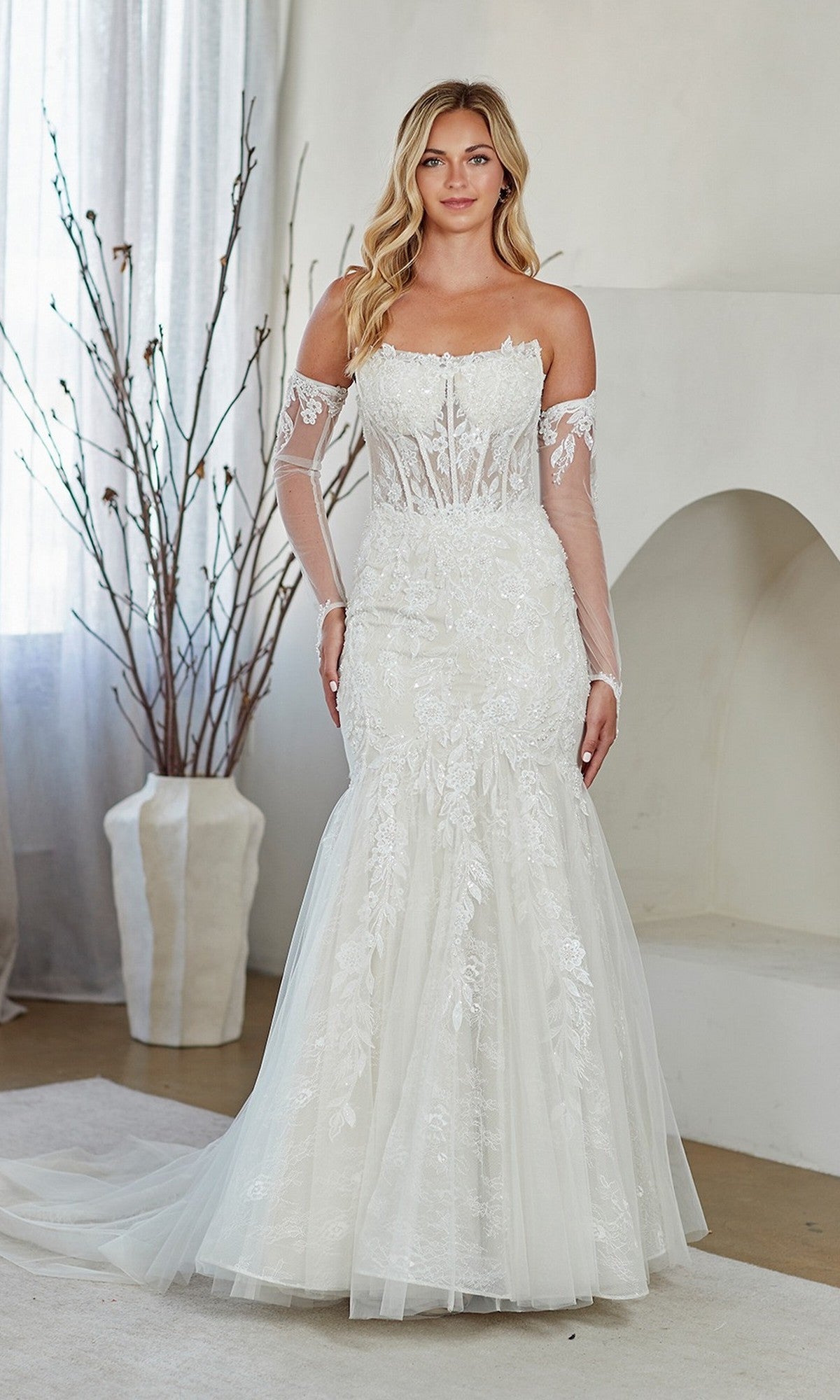 A woman in the Amelia Couture 2155 Long Prom Dress, featuring a strapless corset bodice and lace-embroidered white fabric, stands indoors beside a vase with bare branches.